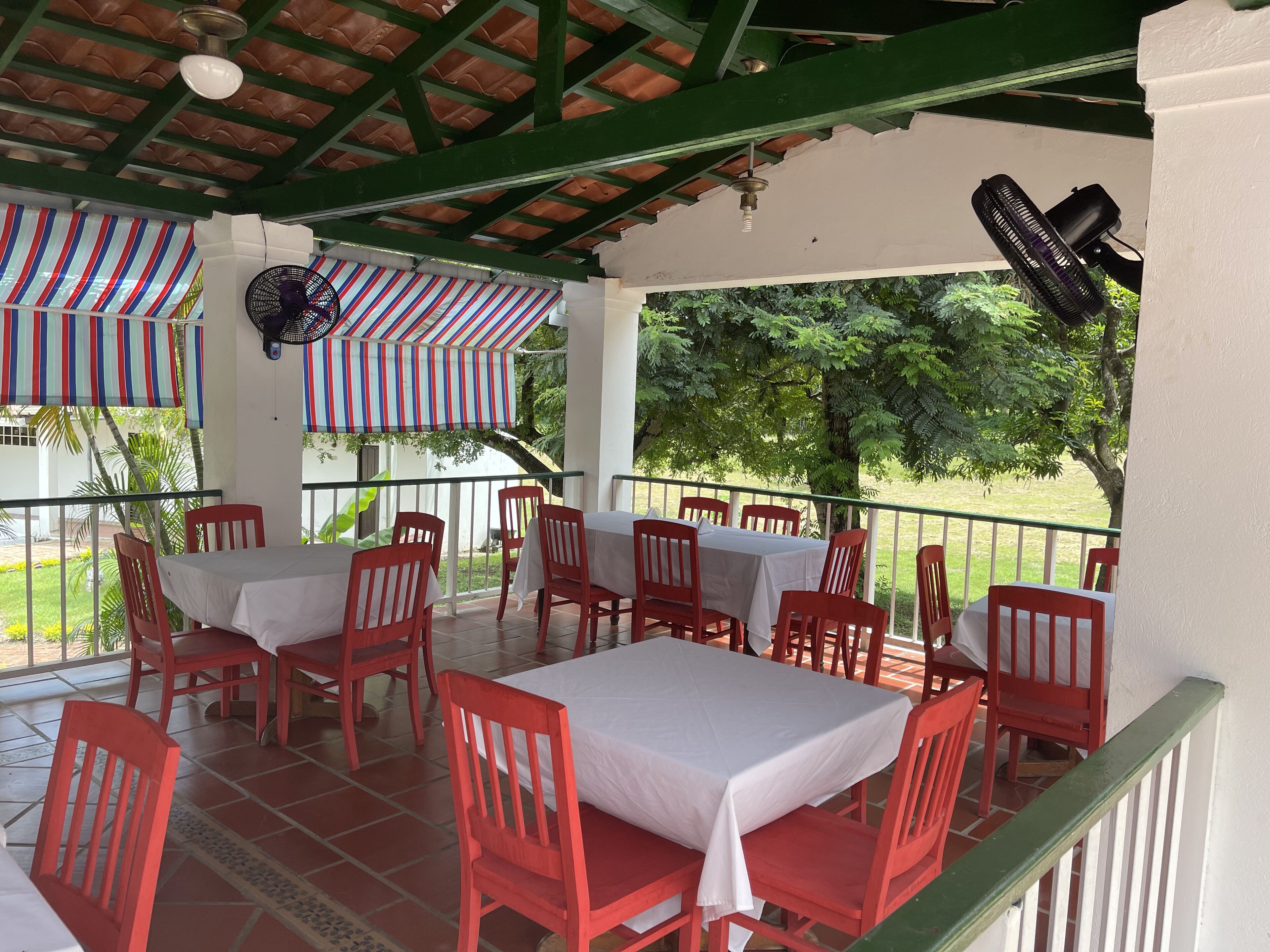 Un área de comedor al aire libre con techo, sillas rojas y mesas blancas, un ventilador de techo y vistas a la vegetación.