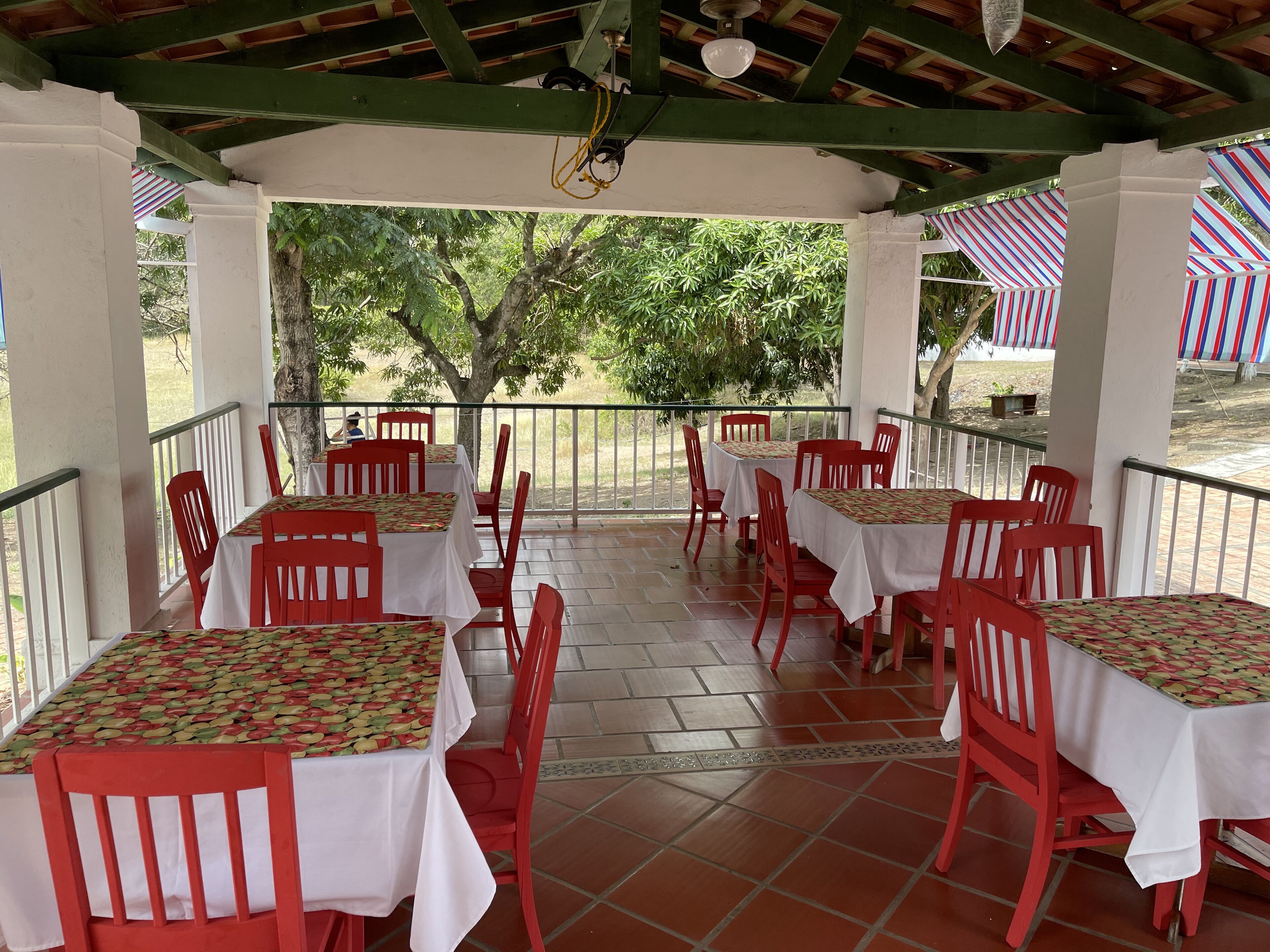 Un área de comedor al aire libre, techada, con mesas y sillas rojas, rodeada por una barandilla y árboles.