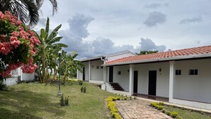 A row of white buildings with red-tiled roofs, a paved walkway, and small landscaped areas with yellow flowers and green shrubs. - HOTEL CAMPESTRE LAS PALOMAS (Tocaima)