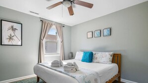 Une chambre à coucher avec un lit, un ventilateur de plafond, une fenêtre avec des rideaux et des œuvres d’art murales.
