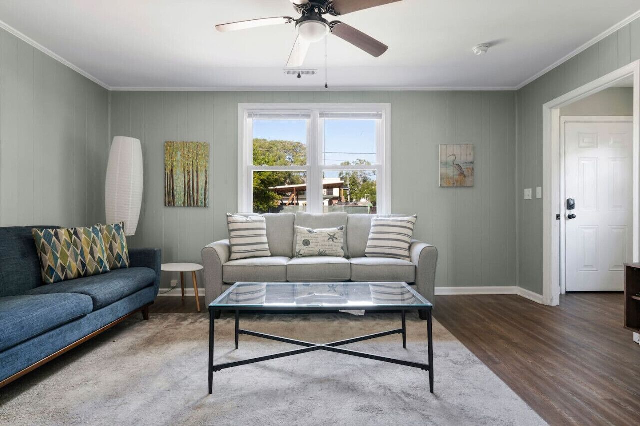 A living room with a sofa, a coffee table, a ceiling fan, and a window with a view of the outdoors.
