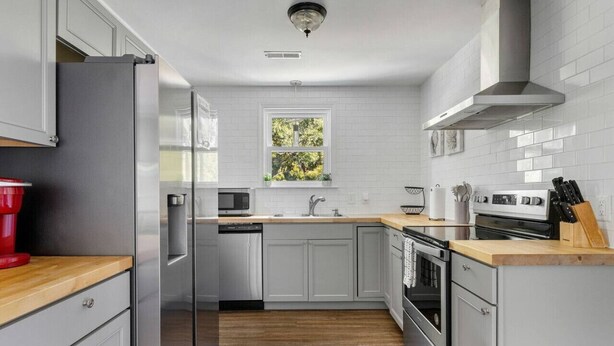 Une cuisine moderne avec des meubles blancs, un plan de travail en bois, des appareils électroménagers en acier inoxydable et une fenêtre donnant sur des arbres.