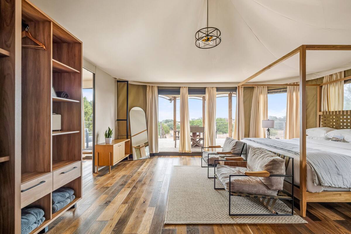 Une chambre avec une armoire en bois, un lit à baldaquin, un coin salon avec un canapé et des fauteuils, une petite table et une grande fenêtre avec des rideaux.