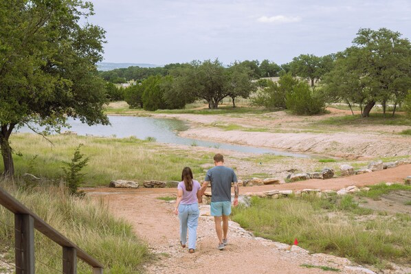 A couple walking on a dirt path beside a river, surrounded by trees and grassy areas.