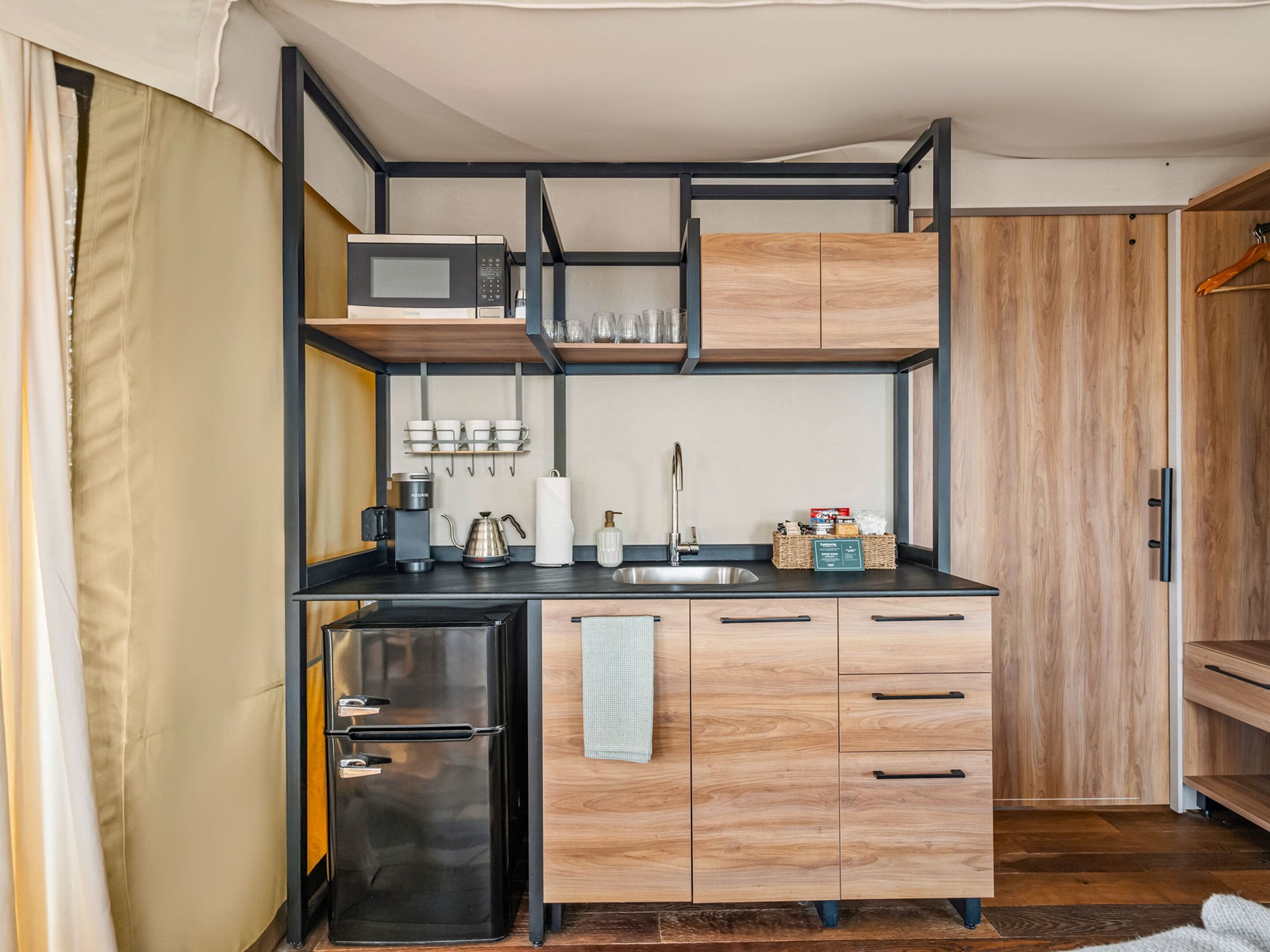 A compact kitchen with a microwave, coffee maker, kettle, and a sink. There are wooden cabinets and a refrigerator.