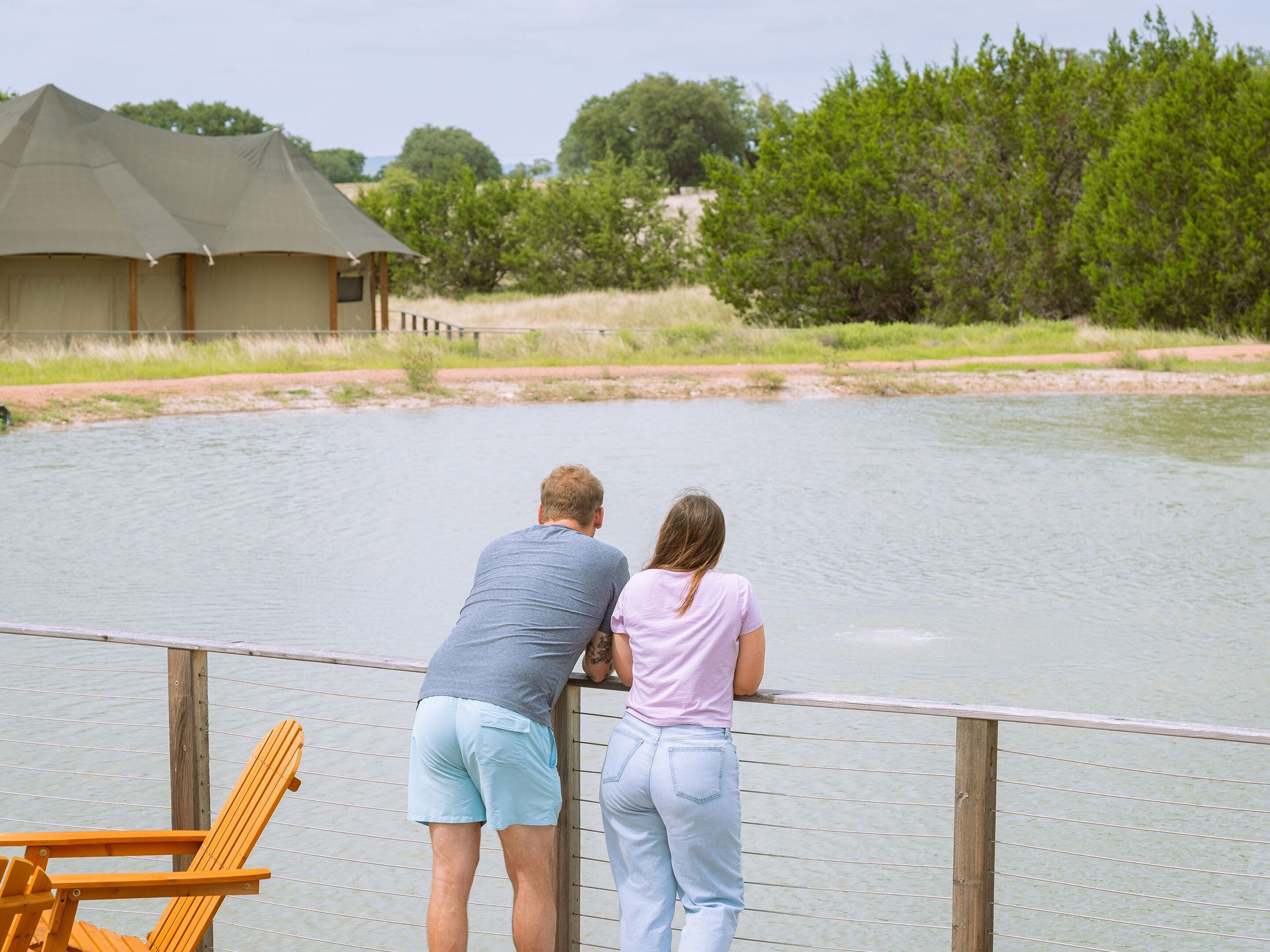 A couple standing on a wooden deck overlooking a body of water, with a tented structure and greenery in the background.