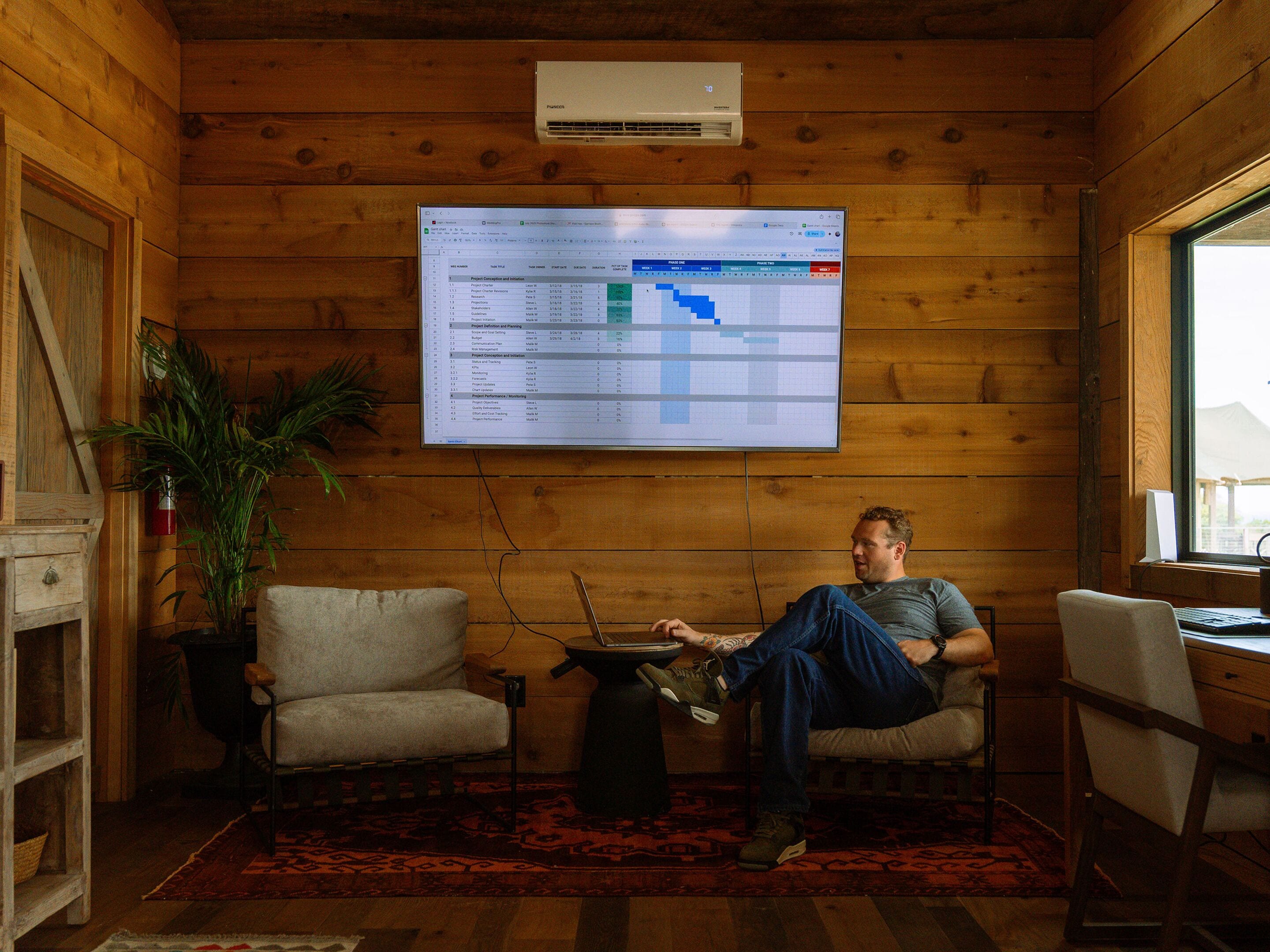 A man sitting in a wooden cabin with a computer monitor displaying a spreadsheet.