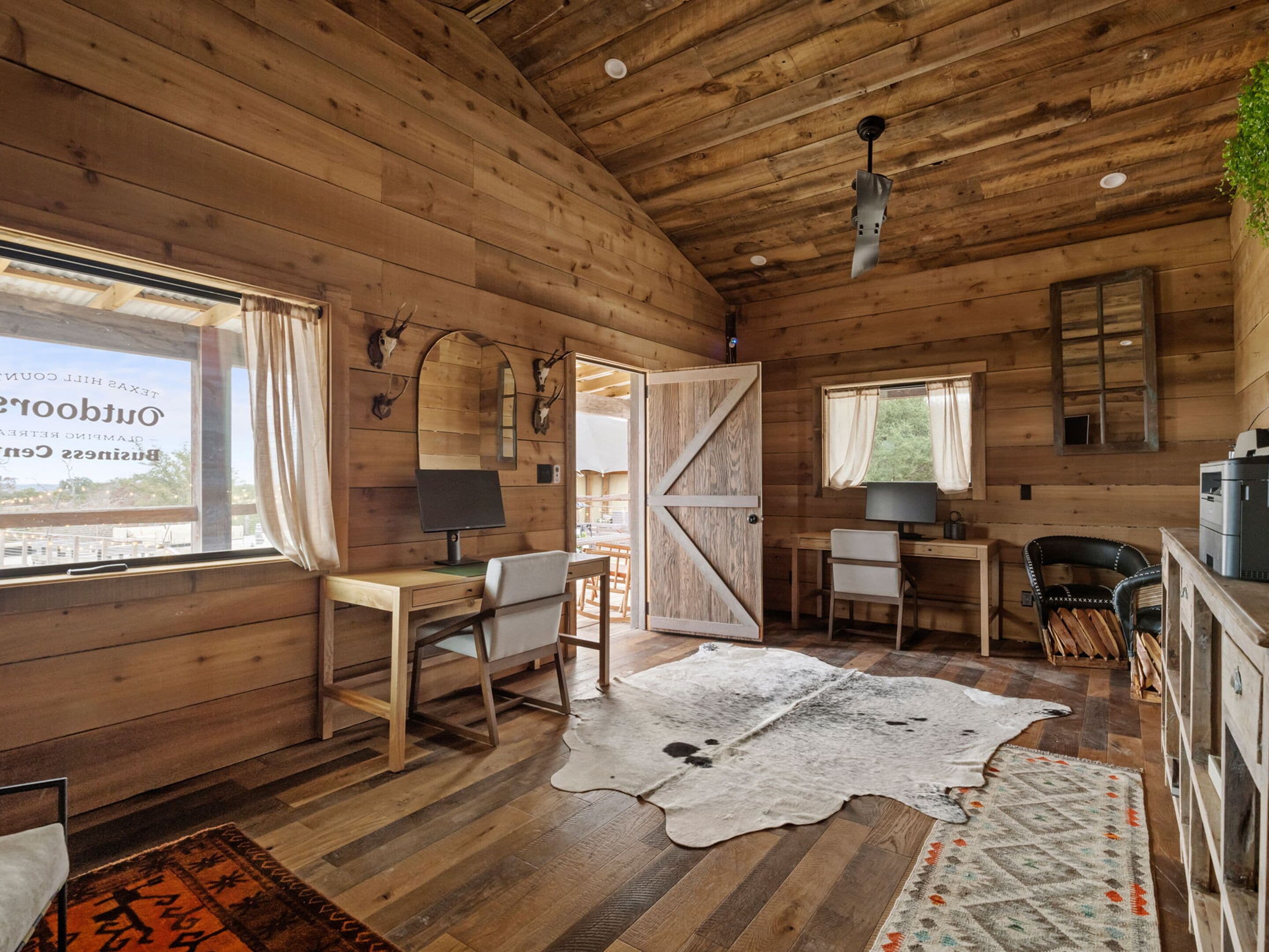 A rustic cabin interior with wooden walls, a desk with a computer, a chair, a large window, and a door.