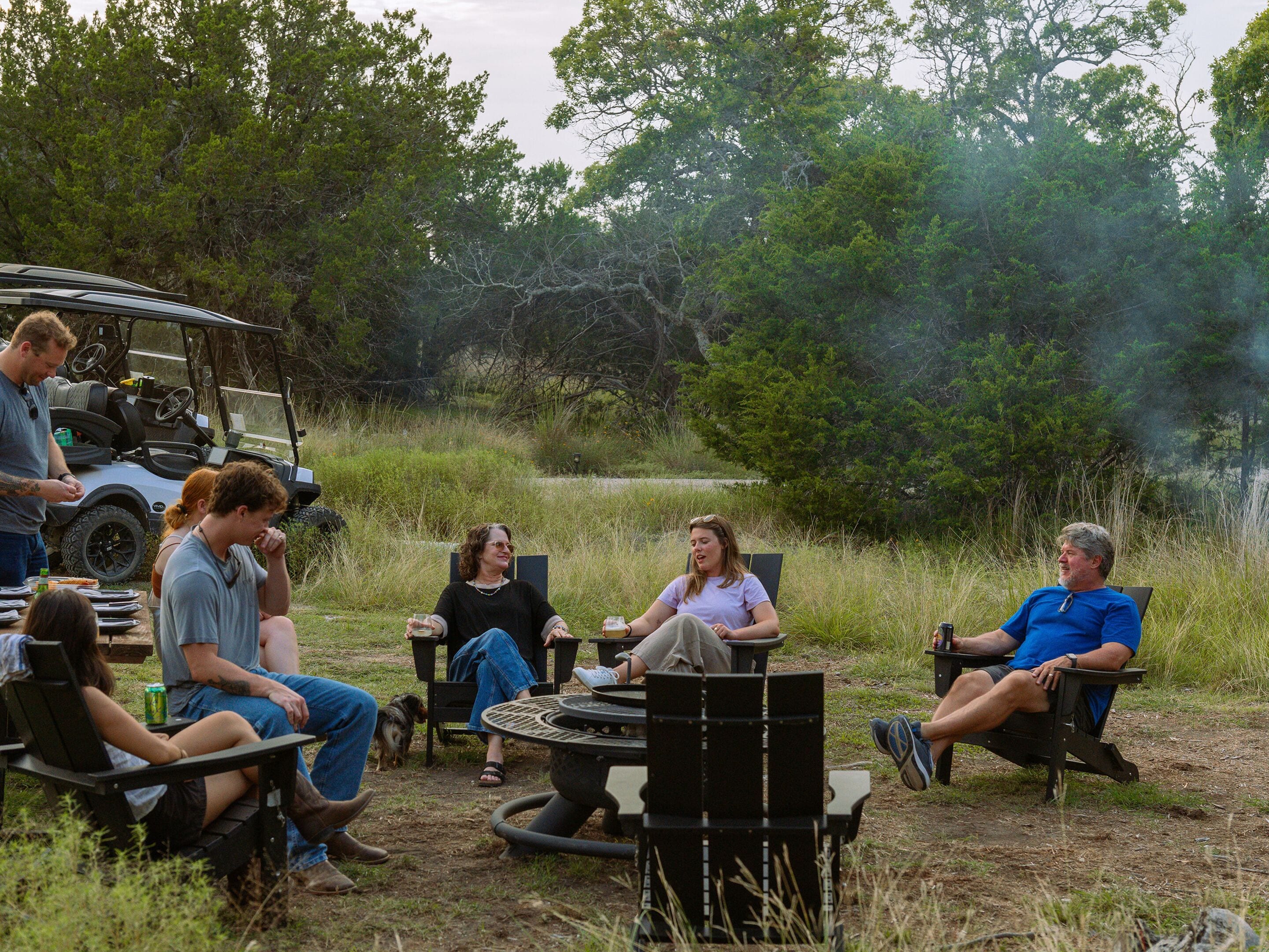 A group of people seated around a circular table outdoors, with a vehicle parked nearby.