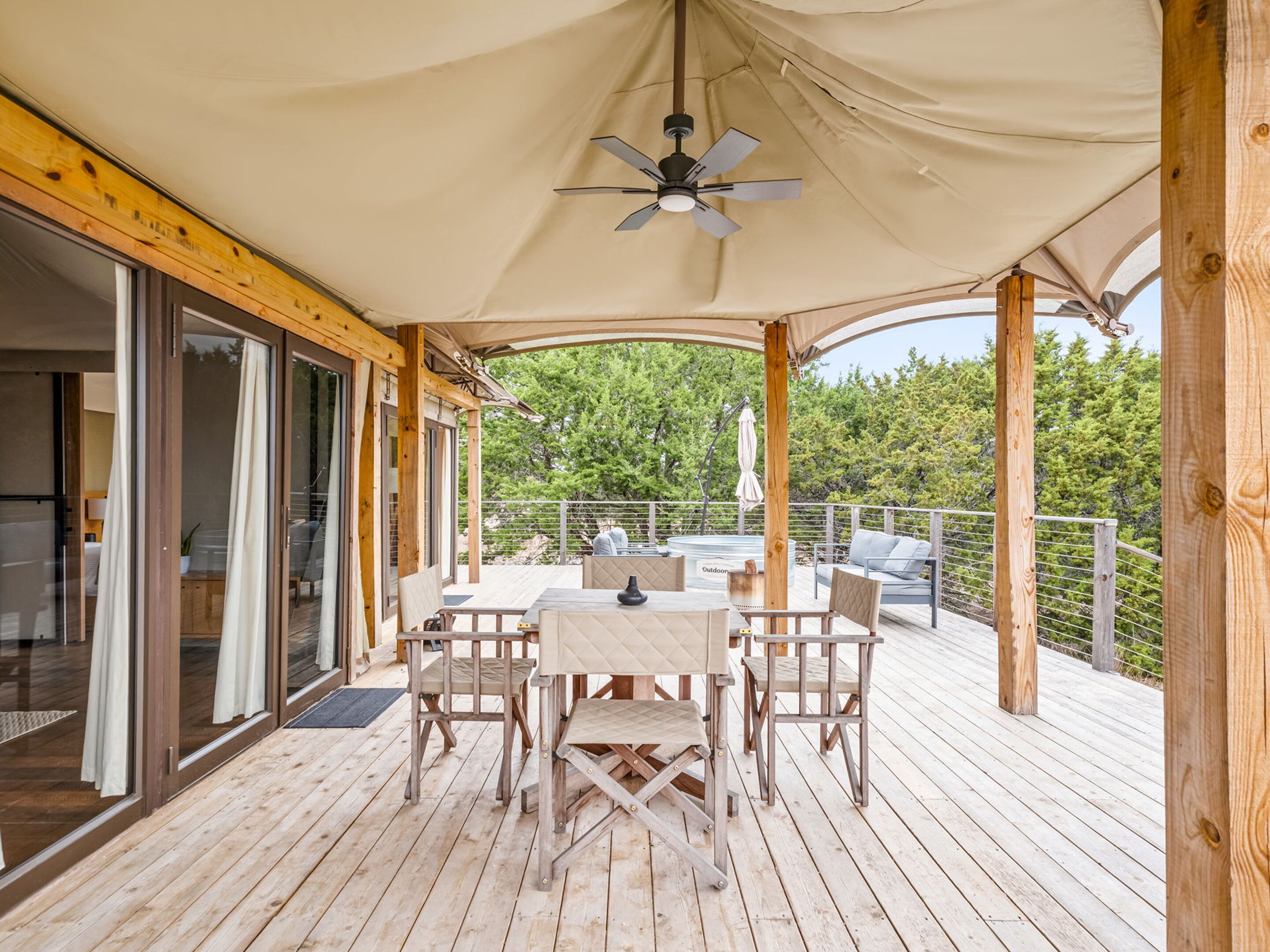 Een houten terras met buitenmeubilair, een plafondventilator en uitzicht op het omliggende bos.