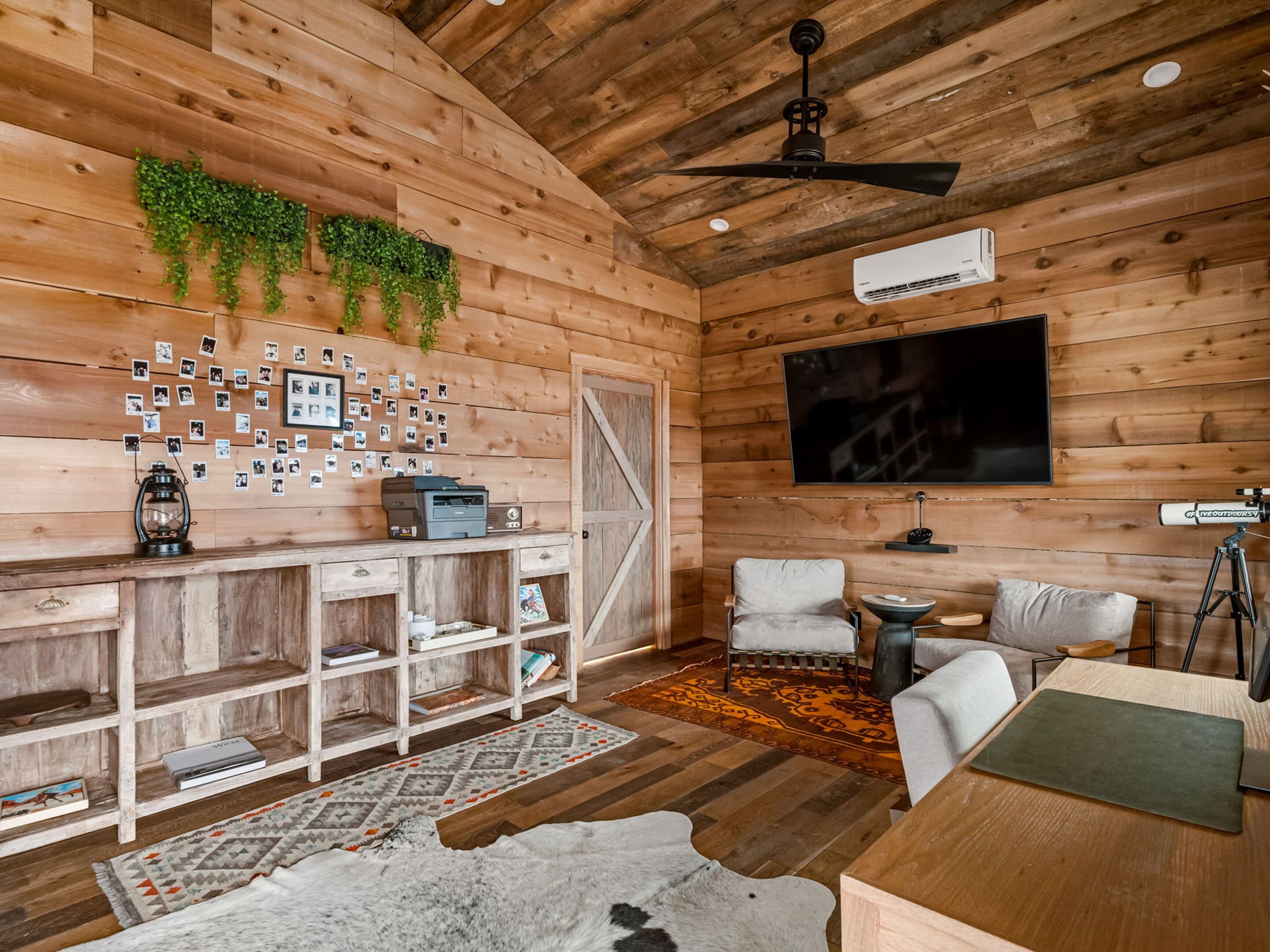 A cozy cabin interior with wooden walls, a mounted TV, a ceiling fan, a bookshelf, and a rustic rug.