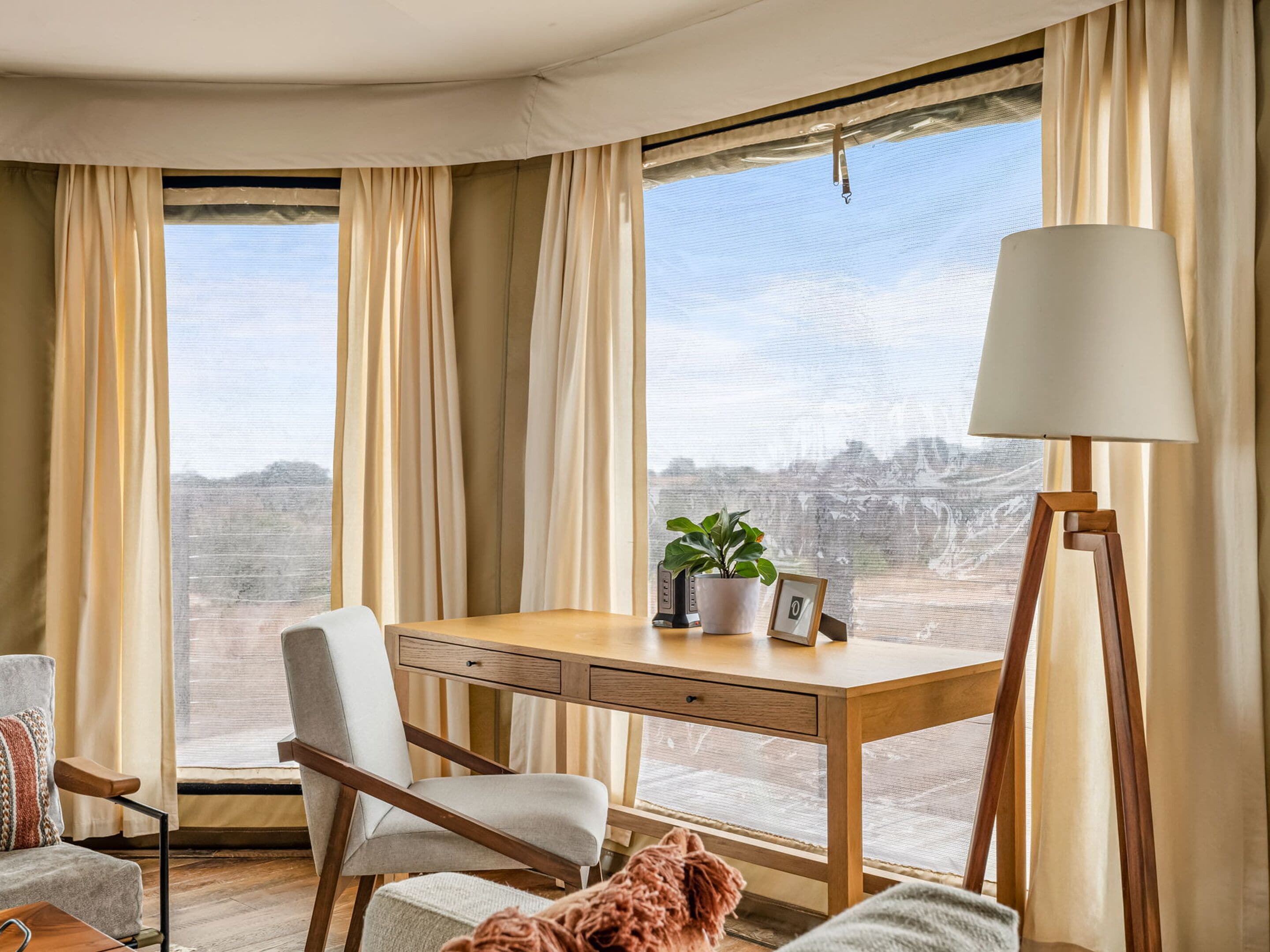 A room with a wooden desk, a chair, a lamp, a potted plant, and a view of a mountainous landscape through large windows.