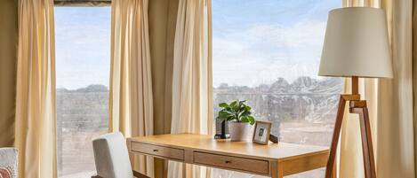 Een kamer met een houten bureau, een stoel, een lamp, een pot met een plant, en uitzicht op een bergachtig landschap door grote ramen.
