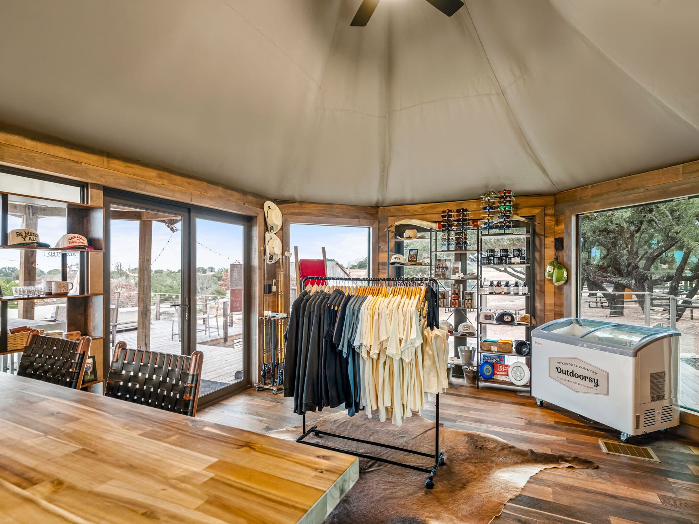 A spacious room with a wooden floor, a large table, and a clothing rack. There is a large window offering a view of the outdoors.