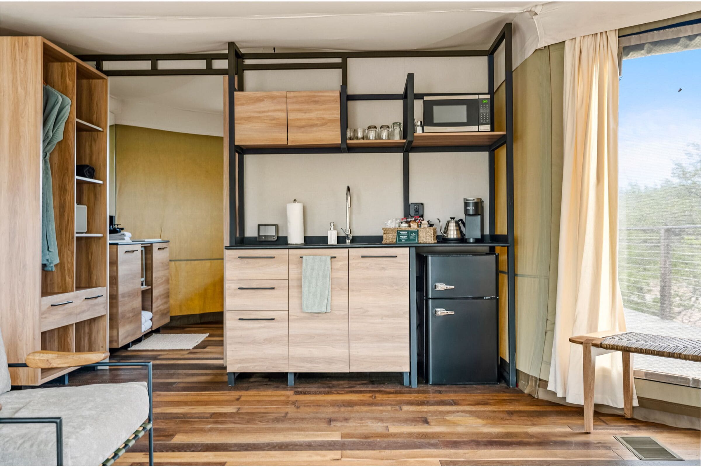 A compact kitchen with wooden cabinets, a microwave, and a small refrigerator. There is a window with curtains and a view of greenery outside.