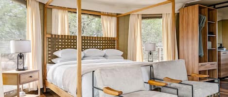 A four-poster bed with a wooden headboard, two bedside tables with lamps, a grey upholstered chair, and a wooden wardrobe.