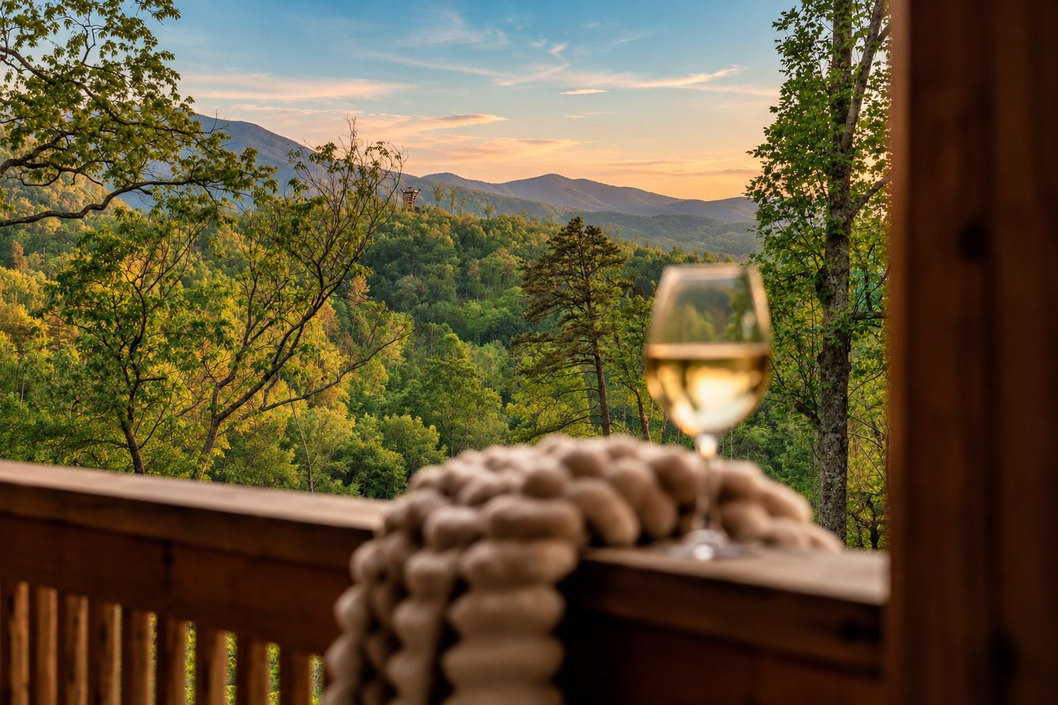 Una terraza de madera con una copa de vino blanco y una manta tejida, con vistas a un paisaje montañoso boscoso.