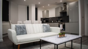 A modern living room with a white sofa, a marble coffee table, and a kitchen area with grey cabinets and a stainless steel range hood.