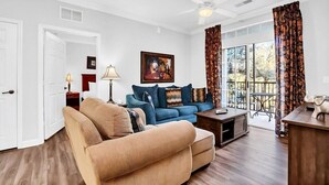 Un salon avec un canapé beige, un canapé bleu, une table basse et un tableau au mur. On y trouve une porte, une lampe et une fenêtre avec des rideaux.