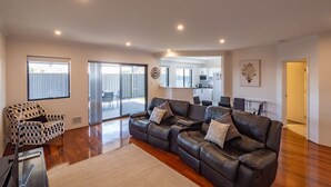 A spacious living room with a large leather sofa, a patterned armchair, and a dining area visible through a sliding glass door.