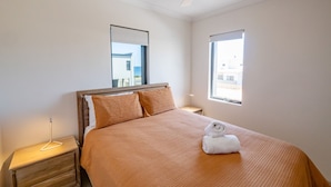 A neatly made bed with a brown bedspread and two pillows, a bedside lamp, and a window with a view of buildings.