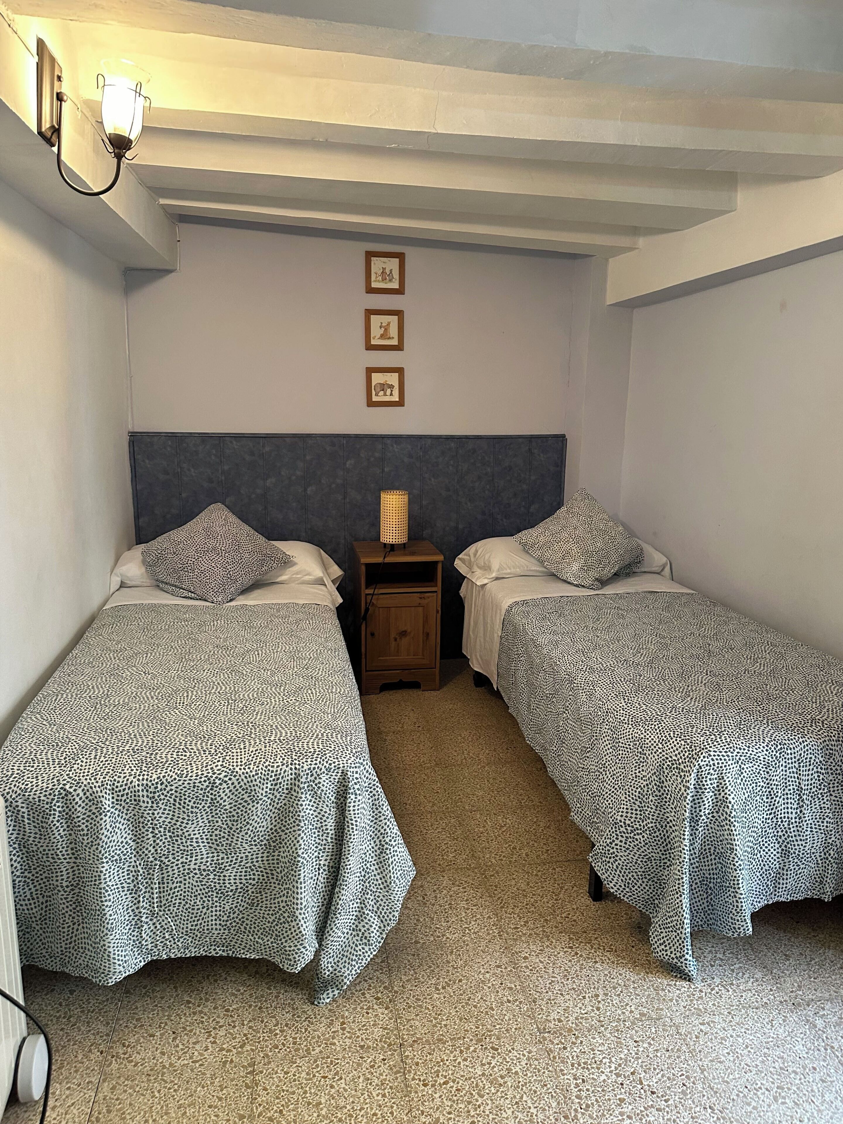 Una habitación con dos camas individuales, una mesita de noche con lámpara, y una pared decorada con tres cuadros.