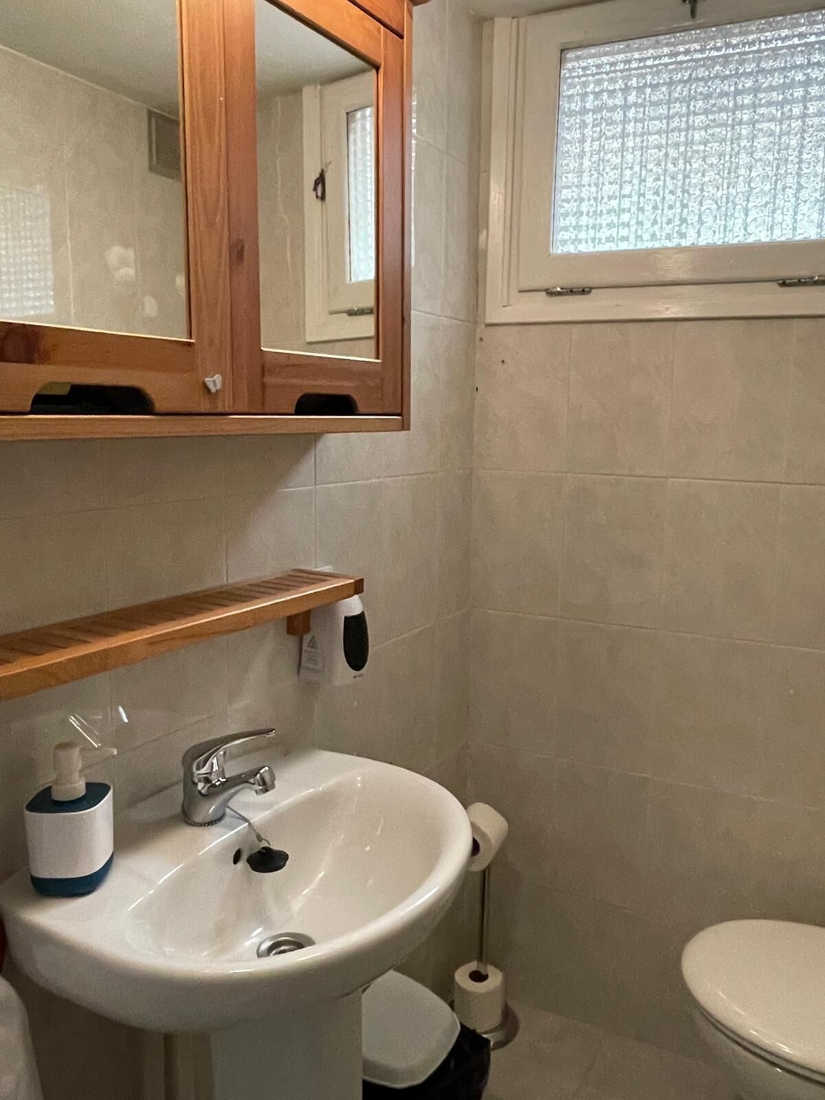 Ett badrum med ett vitt handfat, en spegel, en toalett och ett fönster med mönstrade gardiner.