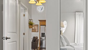 A hallway with a wooden floor, a white door, and a room with a bed and a nightstand. - Cook Cottage (Stockbridge)