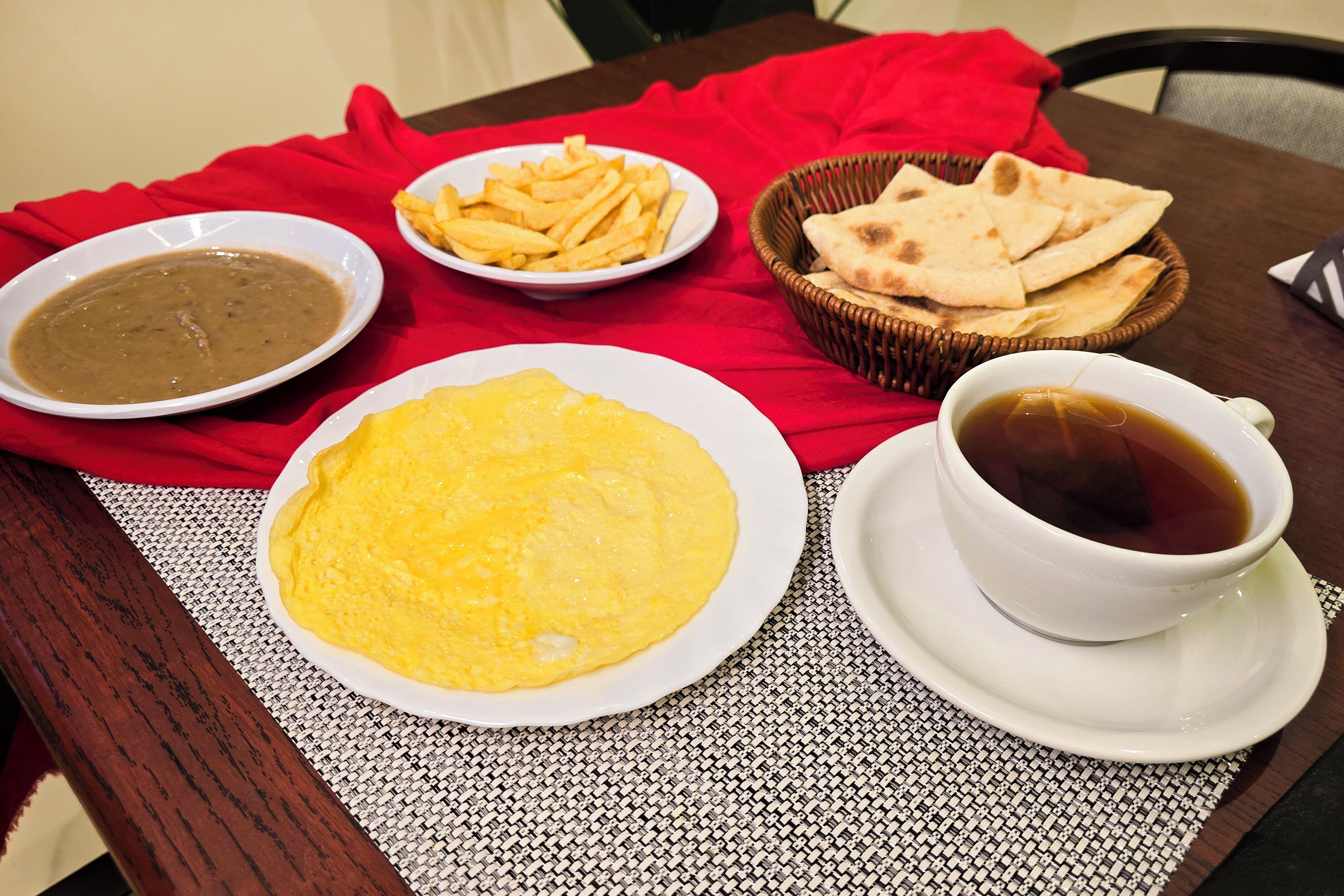 Une table recouverte d’une nappe rouge, une assiette de nourriture jaune, un bol de soupe, un bol de frites, un panier de pain plat et une tasse de thé.