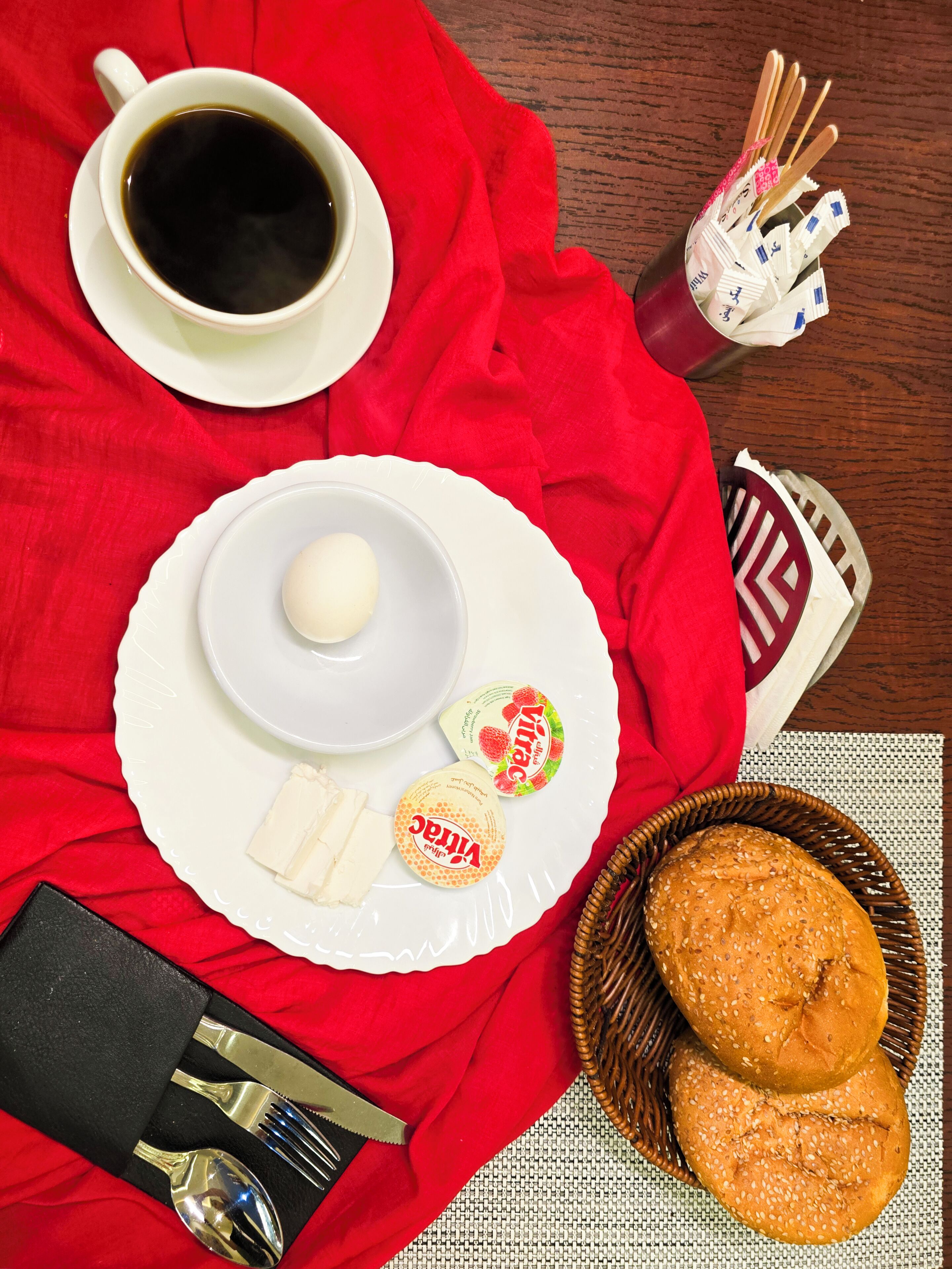 Un desayuno con una taza de café, un plato con un huevo duro, yogur y panecillos sobre un mantel rojo.