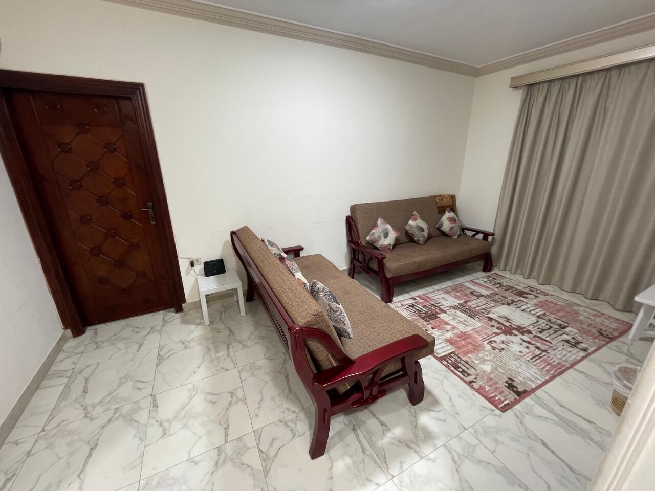 Un salón con sofá, banca, alfombra estampada y una puerta.