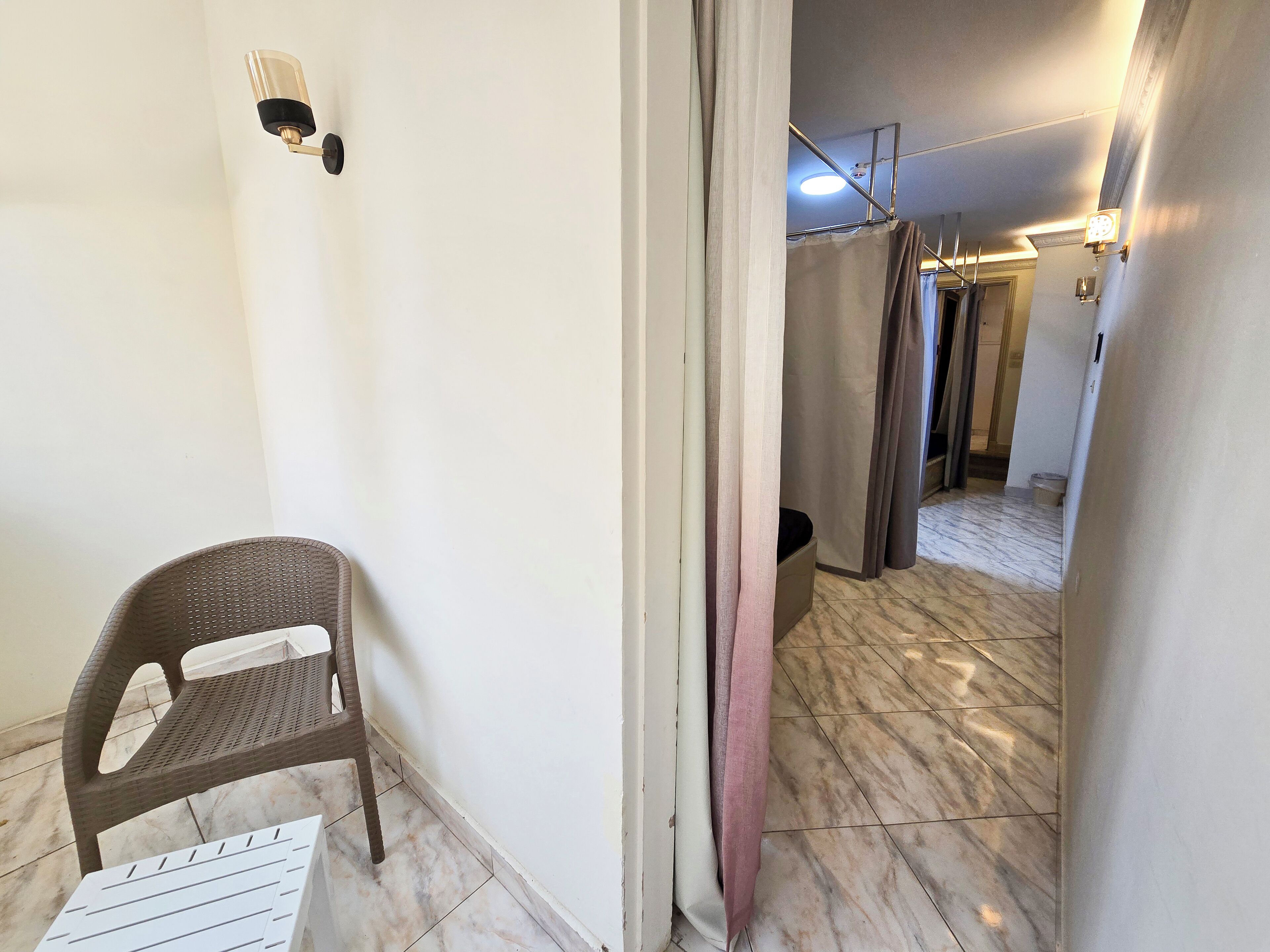 A hallway with a chair, a white wall, and a door with curtains.