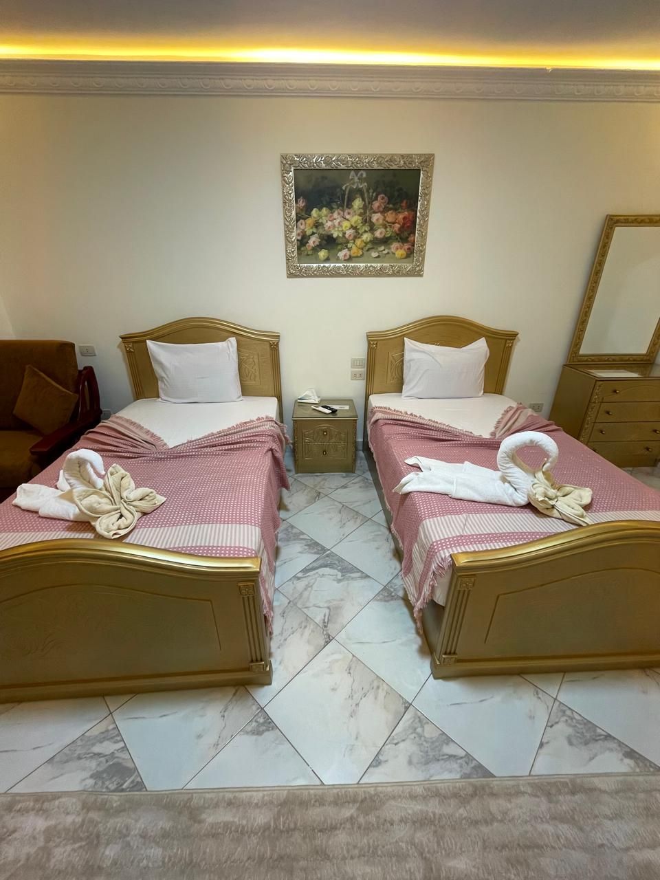 A hotel room with two beds, each with a wooden headboard and a pink bedspread. There is a small table between the beds, a nightstand with a lamp, and a painting on the wall.