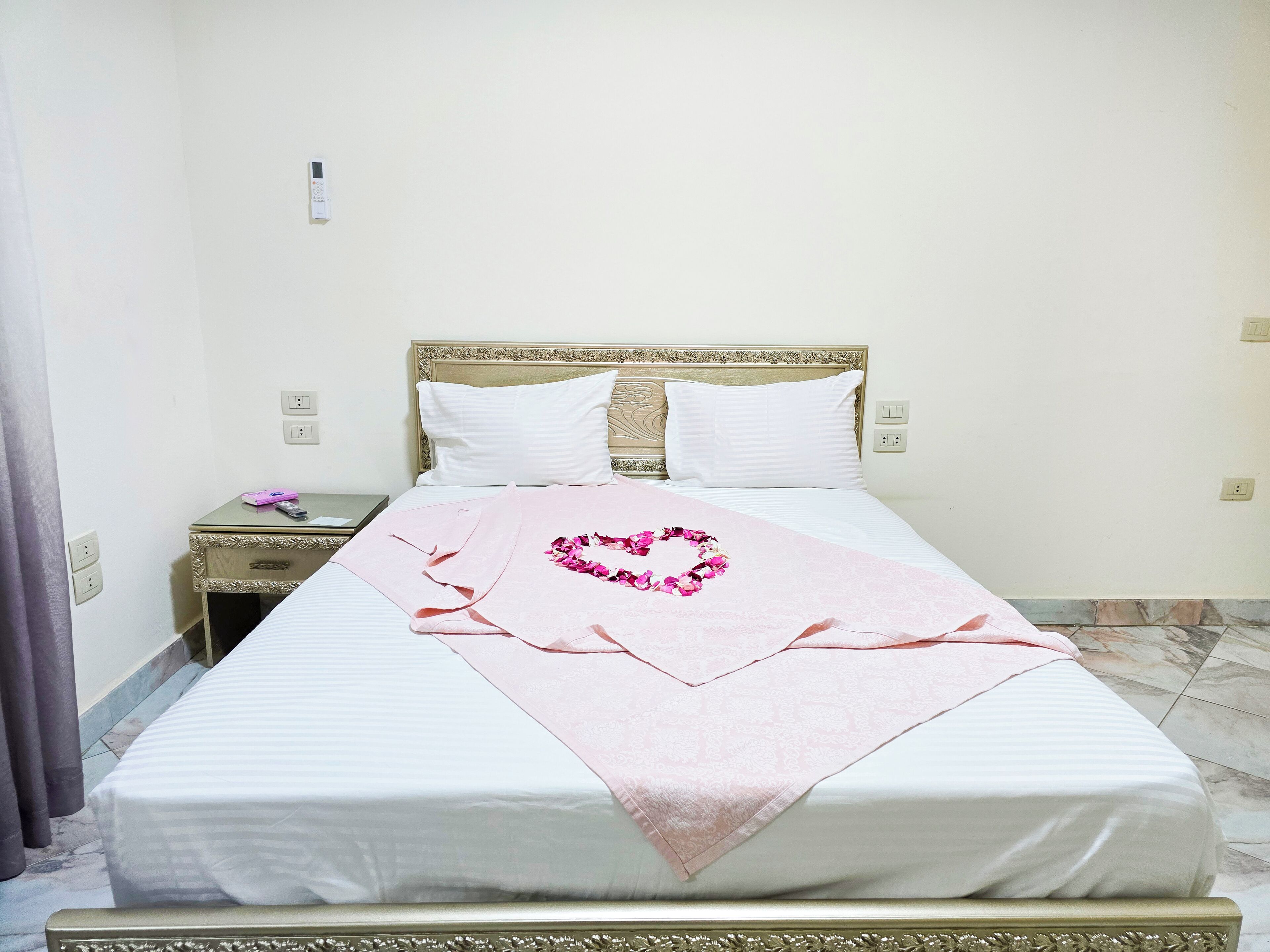 Una cama bien hecha con un adorno en forma de corazón rosa en la colcha, una mesita de noche con un teléfono y un objeto rosa, y una pared lisa con un enchufe.