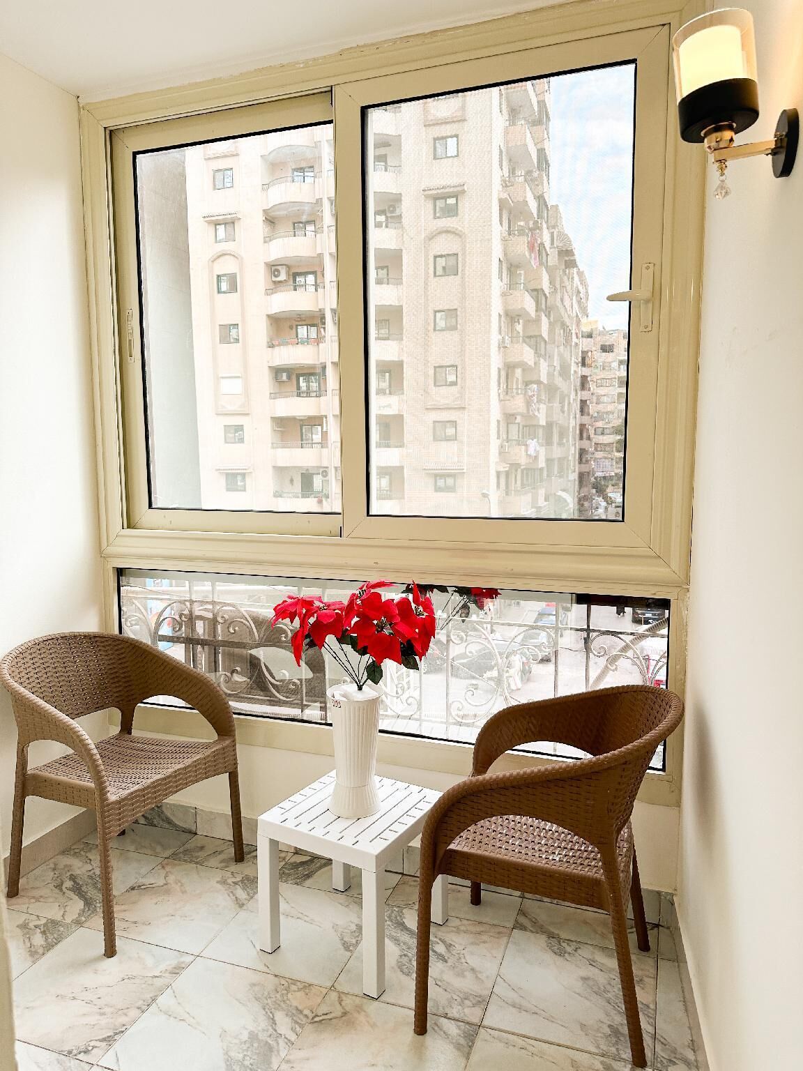 A room with a large window, two wicker chairs, a small white table, and a vase with red flowers.
