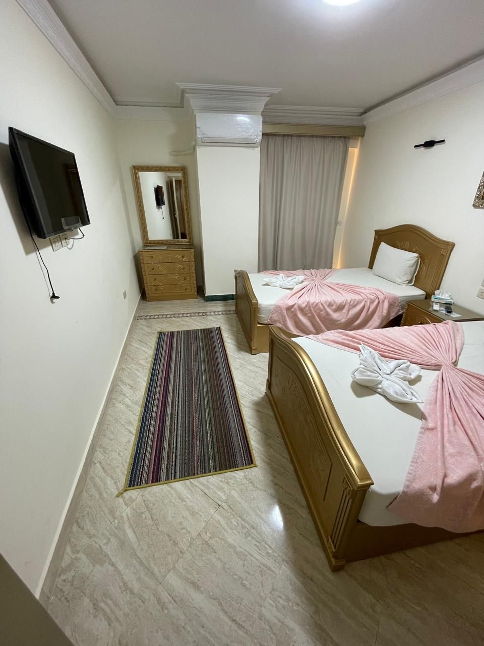 A hotel room with two beds, a TV, a mirror, an air conditioner, and a window with curtains.