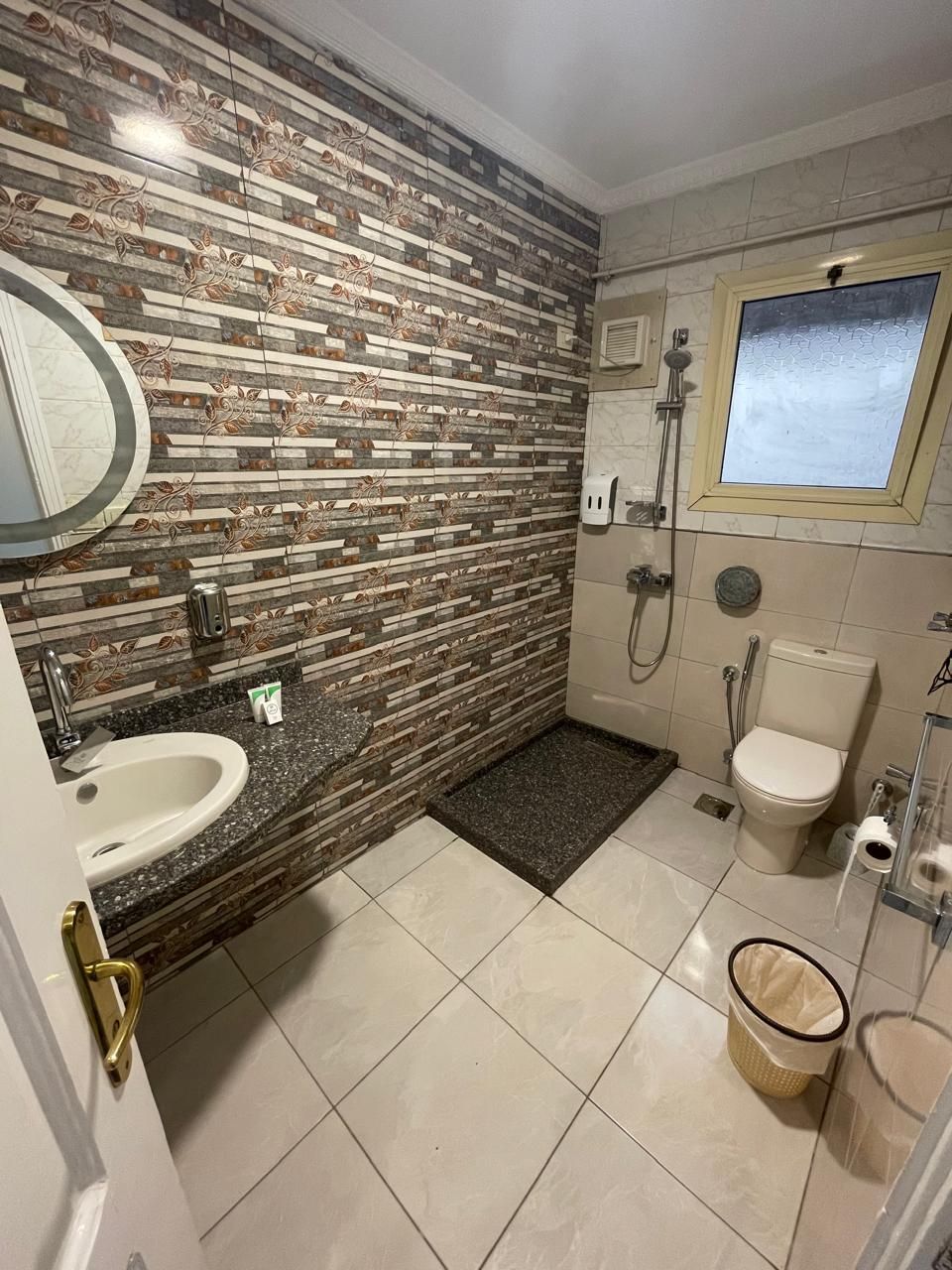 Et badeværelse med flisegulv, en vask med granitbordplade, toilet, bruseområde og et vindue med udsigt.