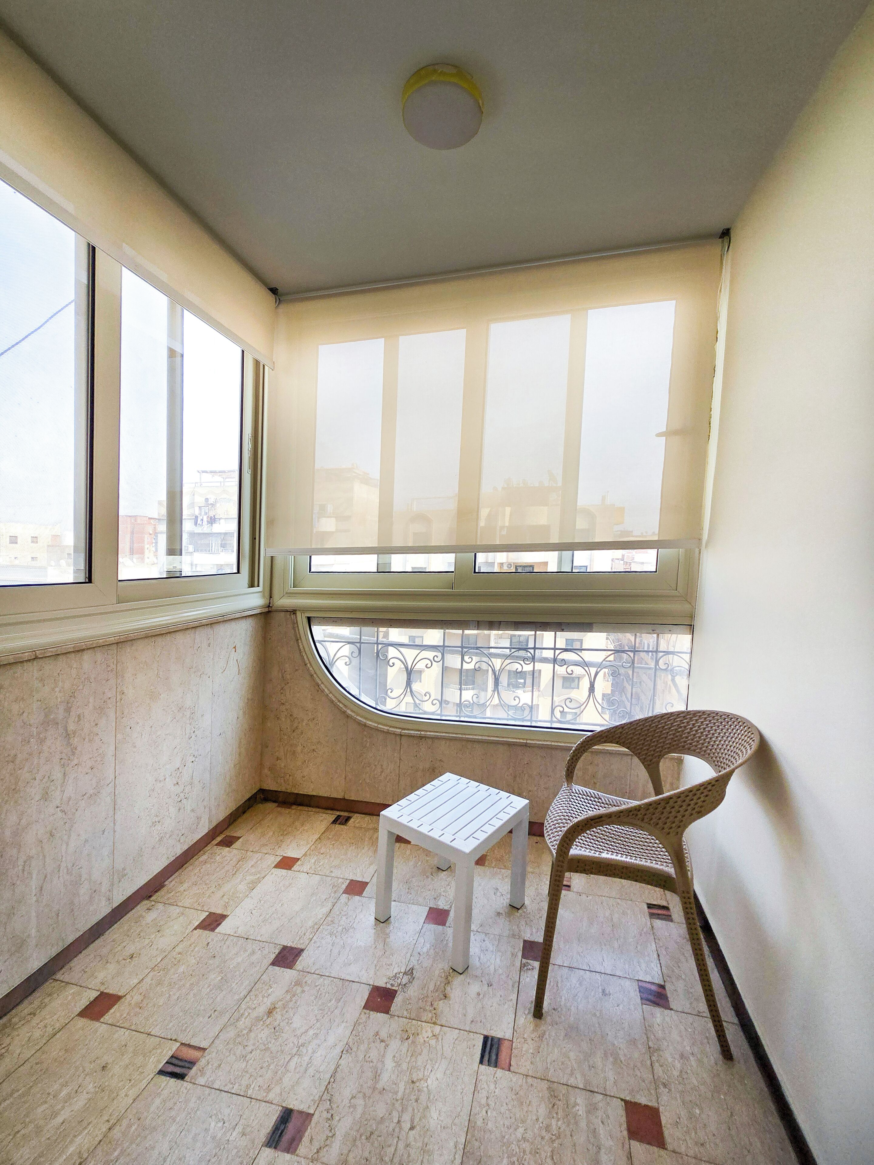 Une pièce d’angle avec une vue dégagée grâce à une grande fenêtre, une petite table blanche et un fauteuil en osier.