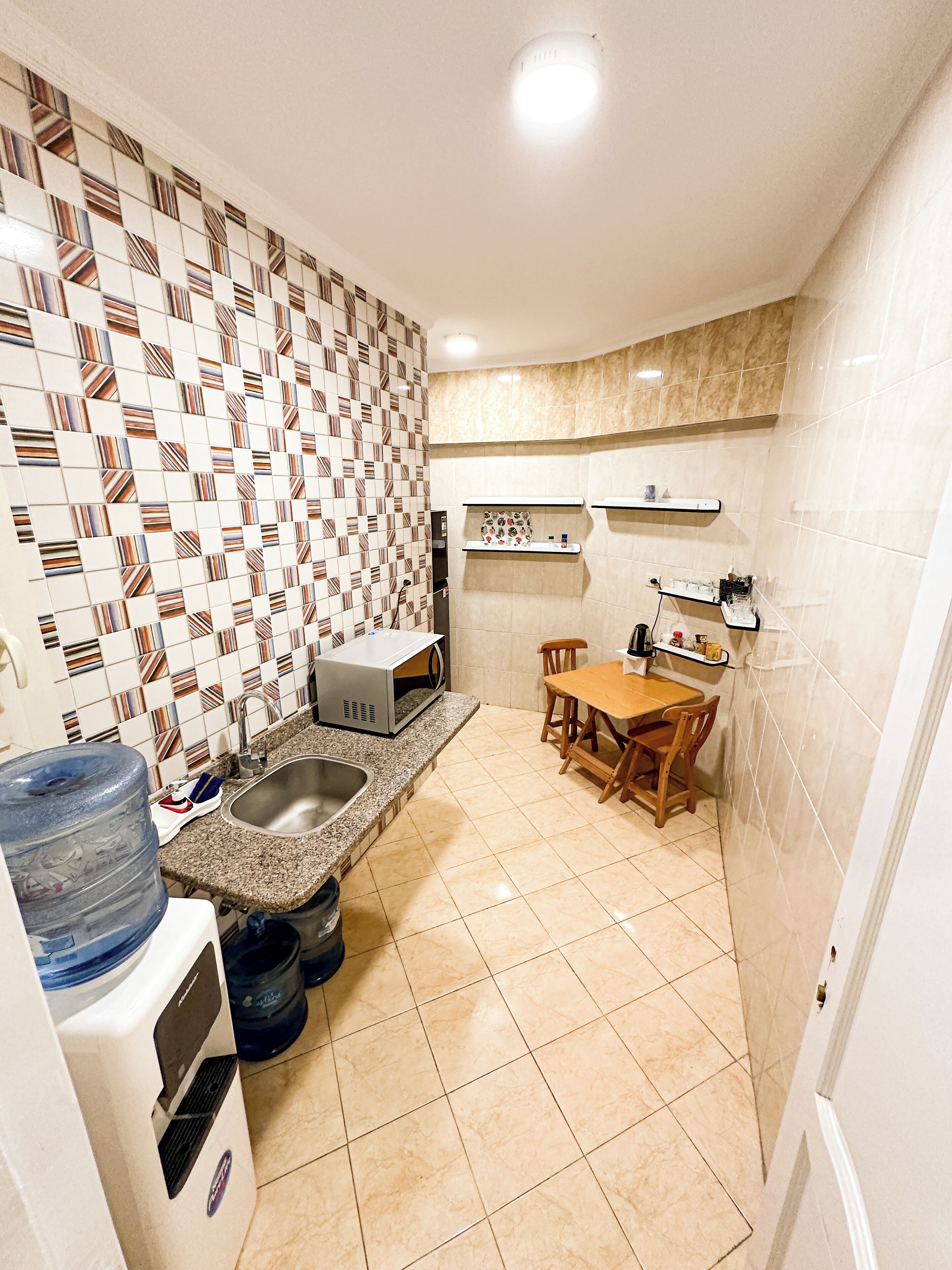 Una cocina compacta con salpicadero de azulejos, un lavamanos, microondas, dispensador de agua y una zona de comedor con mesa y sillas.