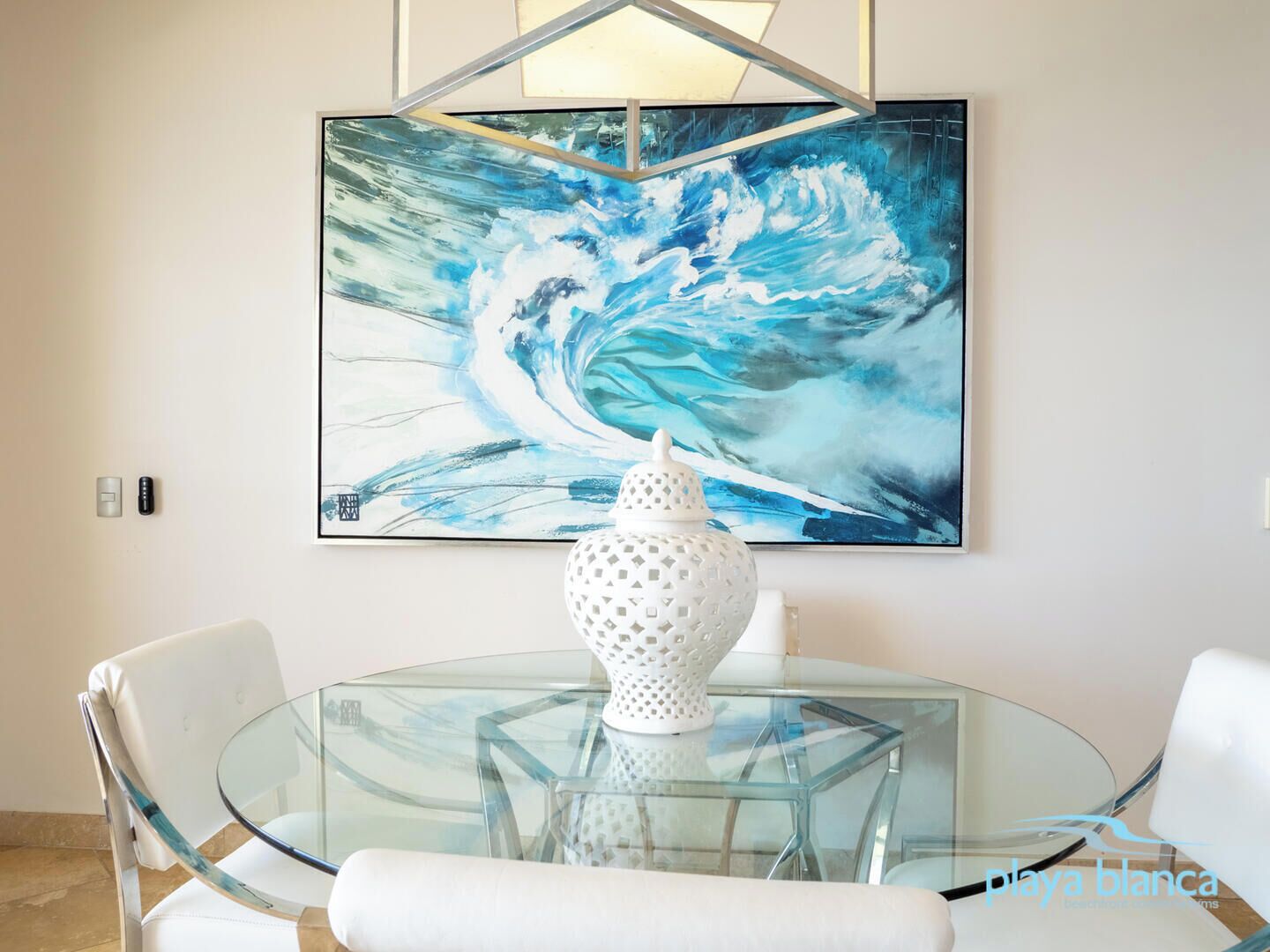 Een eetgedeelte met een glazen tafel, witte stoelen, een witte decoratieve vaas en een groot abstract schilderij aan de muur.