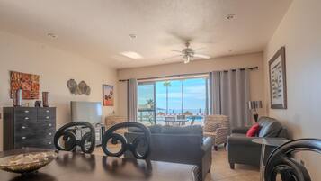Una sala de estar con comedor, ventanas amplias con vista a la playa, ventilador de techo y televisor.