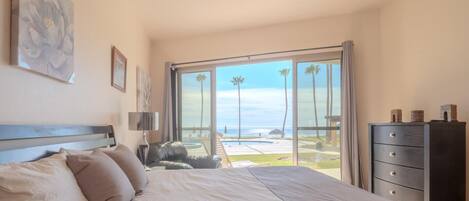 Quarto com uma cama grande, cômoda e vista para a praia e o oceano através de uma porta de vidro deslizante.