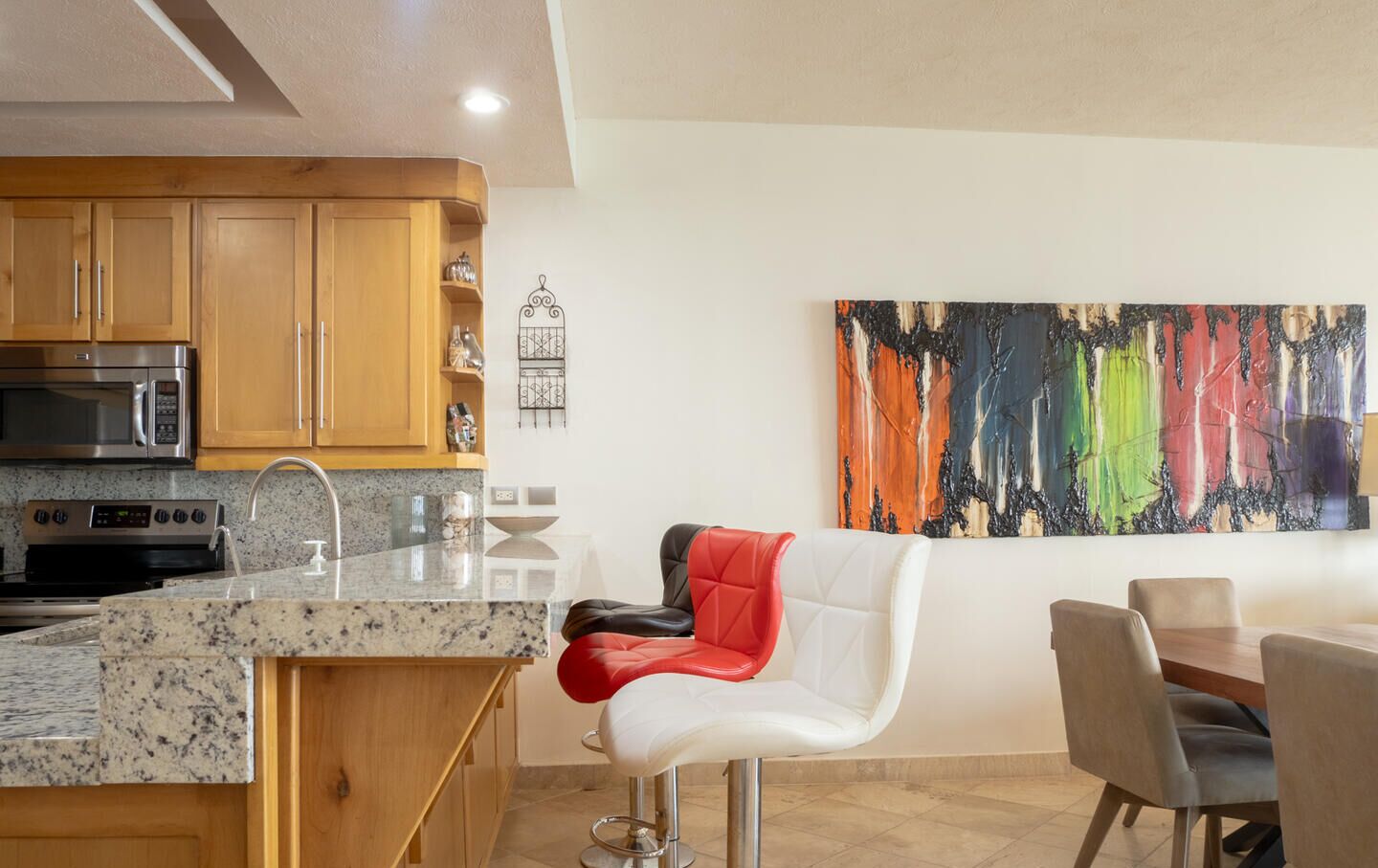 Une cuisine moderne avec des comptoirs en granit, des appareils en acier inoxydable et des armoires en bois. On y trouve un coin repas avec une chaise blanche et une chaise rembourrée rouge. Un tableau abstrait aux couleurs vives est accroché au mur.