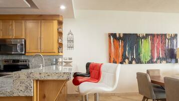 Une cuisine moderne avec des comptoirs en granit, des appareils en acier inoxydable et des armoires en bois. On y trouve un coin repas avec une chaise blanche et une chaise rembourrée rouge. Un tableau abstrait aux couleurs vives est accroché au mur.