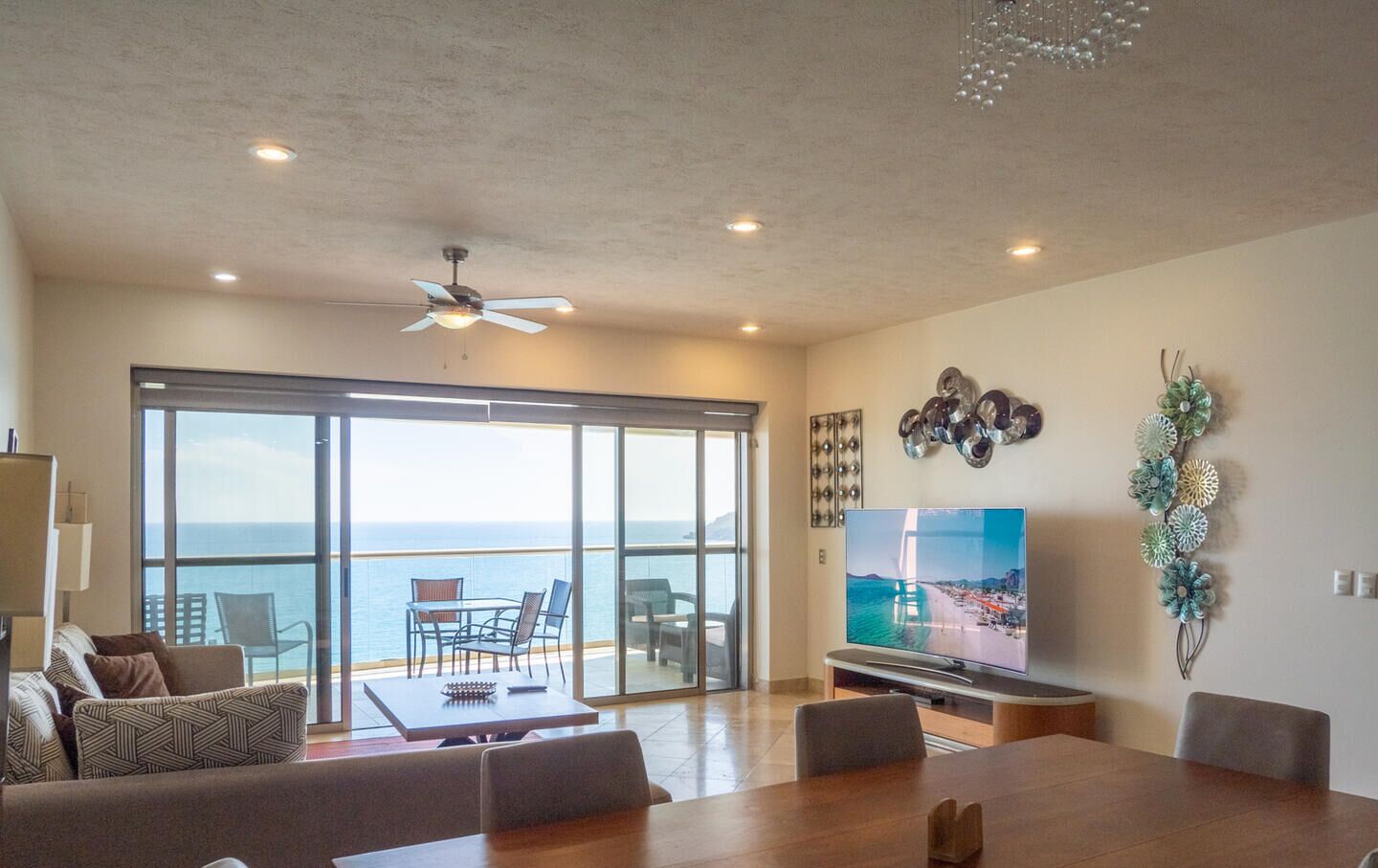 A spacious living room with a large TV, a dining area, and a balcony with a view of the sea.
