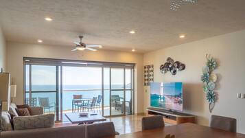 A spacious living room with a large TV, a dining area, and a balcony with a view of the sea.