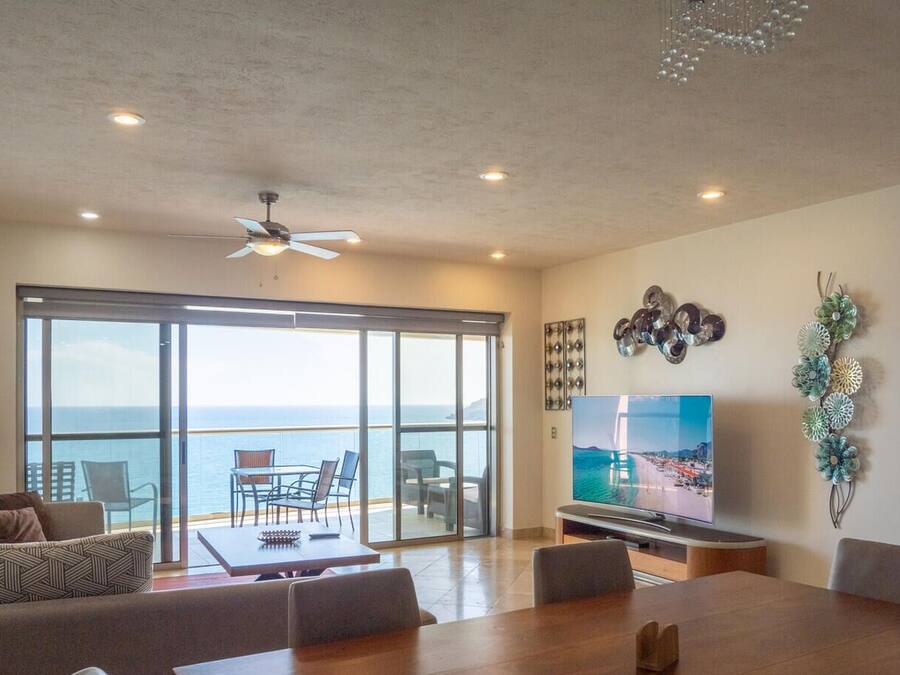A spacious living room with a large TV, a dining area, and a balcony with a view of the sea.