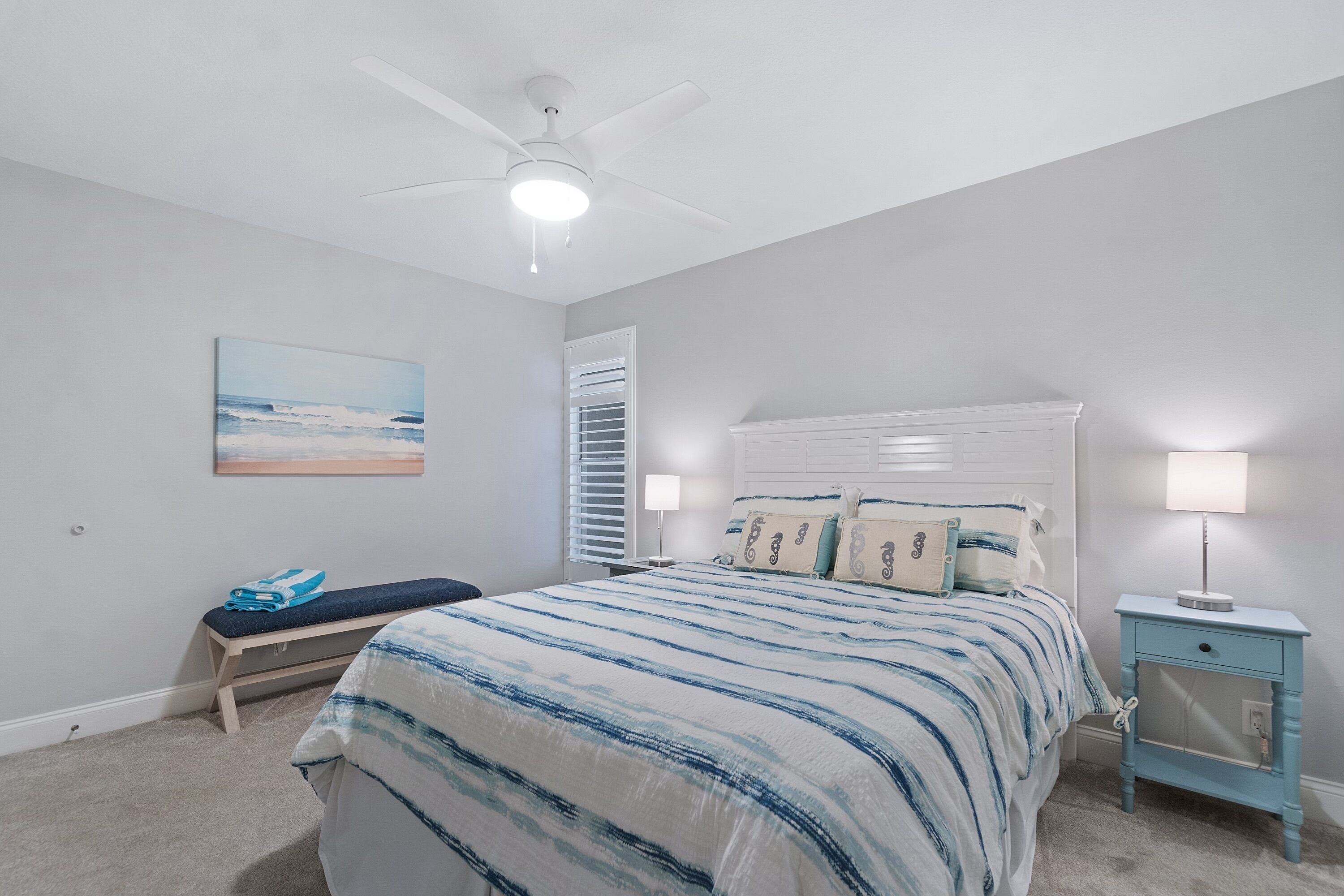 A bedroom with a bed, a bench, a nightstand, a ceiling fan, and a painting on the wall.