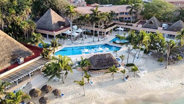 Eine Vogelperspektive zeigt einen Ferienort mit einem zentralen Pool, Palmen und Strandbereichen.