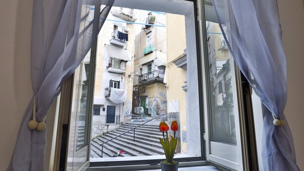 A window with sheer curtains, a potted plant with red flowers, and a view of a narrow street with buildings. - GuestHost - Porta Medina Apartment (Napoli)